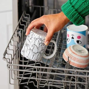 May include: A white mug with a black, abstract wave pattern is placed in a dishwasher. Other dishware includes a blue and white mug with a cartoon face and a small bowl with a red dotted pattern.