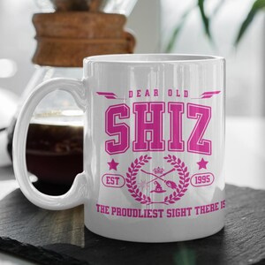 May include: White ceramic mug with the text "DEAR OLD SHIZ" in pink, along with a crest design and the text "EST 1995". The mug has a large handle and is set against a blurred background.