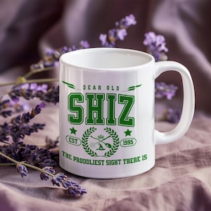 May include: White ceramic mug with a green design that reads "DEAR OLD SHIZ" in a bold, collegiate-style font. The mug also features a crest with the text "EST 1995" and the phrase "THE PROUDLIEST SIGHT THERE IS".