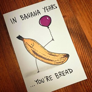 May include: A greeting card with a banana illustration holding a purple balloon. The text reads "In Banana Years... You're Bread". The banana is yellow and brown with a sad face. The background is a wooden surface.