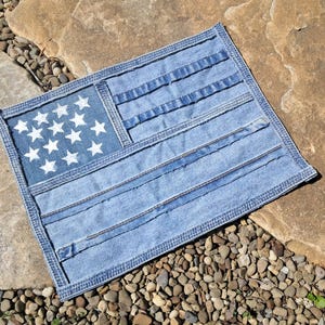 May include: A denim flag with a patchwork design. The flag features a blue field with white stars and blue and white stripes, all made from repurposed denim. The flag is rectangular and has a stitched border.