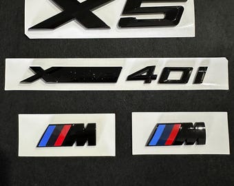 May include: Black car emblems, including "X5", "X4 40i", and two "M" badges with blue, red, and black stripes. The emblems are on white backing paper, ready for application. Automotive accessories.
