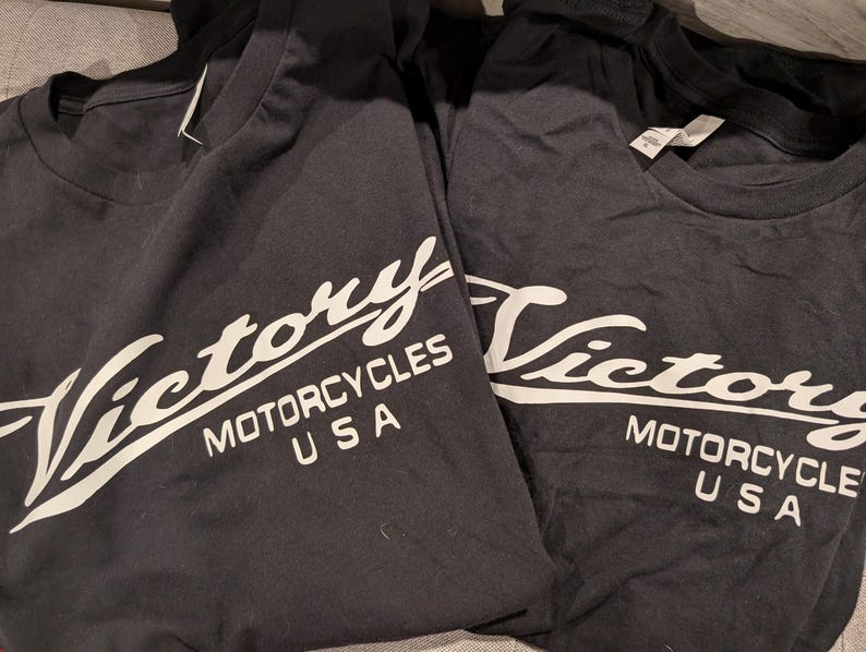 May include: Two black t-shirts with a white "Victory Motorcycles USA" logo. The logo is in a stylized script font.