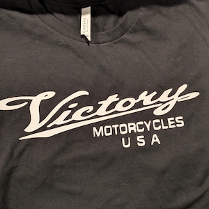 May include: Black t-shirt with a white graphic of the Victory Motorcycles logo. The logo features the word "Victory" in a stylized script font with the words "Motorcycles USA" underneath.