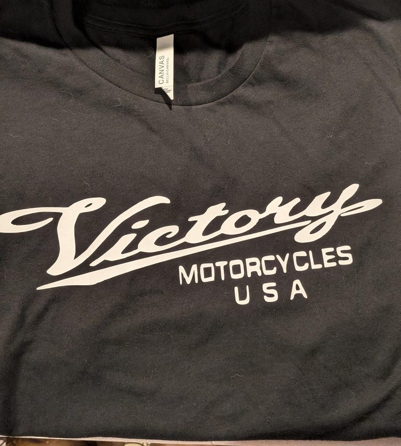 May include: Black t-shirt with a white graphic of the Victory Motorcycles logo. The logo features the word "Victory" in a stylized script font with the words "Motorcycles USA" underneath.