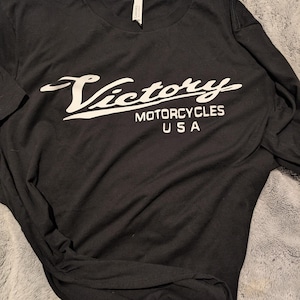 May include: Black t-shirt with a white graphic of the Victory Motorcycles logo. The logo is a stylized script font with the words "Victory Motorcycles USA".
