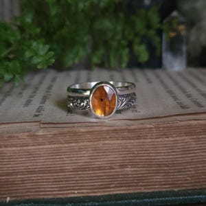 May include: A silver ring featuring an oval-shaped, faceted orange gemstone set in the center. The band of the ring displays intricate, embossed scrollwork patterns along its sides, providing a detailed texture to the metallic surface.