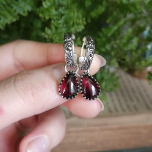 May include: A pair of silver hoop earrings featuring intricate floral engravings on the metal bands. Each hoop suspends a deep red, pear-shaped gemstone charm with a faceted surface, secured by a small silver setting with a beaded edge.