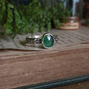 May include: A silver ring featuring a polished, oval-shaped green gemstone set in a smooth bezel. The band is crafted from silver with a detailed, embossed pattern of small circles and cross-like shapes along the outer surface of the metal.