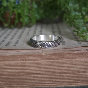 May include: A silver ring featuring a continuous embossed pattern of stylized leaves around the outer band. The metal has a polished interior and a darkened, oxidized finish within the recessed areas of the leaf design on the exterior surface.