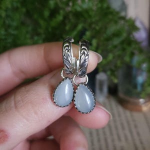 May include: A pair of silver hoop earrings featuring an intricate engraved leaf pattern on the hoops and dangling teardrop-shaped, pale blue cabochon stones set in scalloped silver bezels, creating a cohesive and detailed jewelry piece.