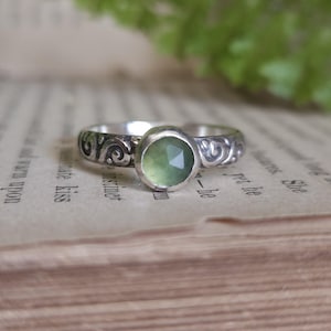 May include: A silver ring with a light green, faceted gemstone. The band features an ornate, swirling design. The ring is set against a backdrop of an open book.