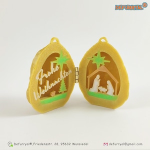 Può includere: Un ornamento dorato con cerniera con una scena della Natività di Natale su un lato e le parole "Frohe Weihnachten" sull'altro.