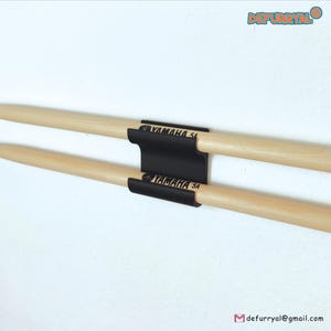 Pode incluir: Duas baquetas de madeira presas por um suporte preto. As baquetas são castanho claro com pontas arredondadas. O suporte tem o logotipo "YAMAHA 5A". O fundo é branco.