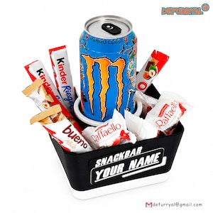 Könnte beinhalten: Ein schwarz-weißer Snackbehälter mit der Aufschrift SNACKBAR YOUR NAME, gefüllt mit einer blauen Monster Mango Loco Energy-Drink-Dose, zwei Kinder Bueno Riegeln, zwei Kinder Riegeln und zwei Packungen Raffaello Kokos-Mandel-Konfekt.