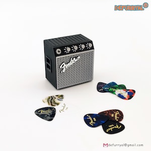 以下が含まれることがあります： 黒いミニチュアギターアンプ。銀色のスピーカーグリルとFenderのロゴがあります。アンプの周りには、さまざまなデザインとFenderのロゴが入った、カラフルなギターピックがいくつか散らばっています。