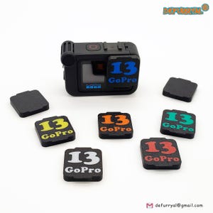 Könnte beinhalten: Eine schwarze GoPro-Kamera mit einem blauen "13 GoPro"-Aufkleber. Mehrere schwarze quadratische Zubehörteile und zusätzliche Aufkleber mit dem Text "13 GoPro" in verschiedenen Farben, darunter Gelb, Orange, Grün, Rot und Weiß, sind um die Kamera herum verstreut.