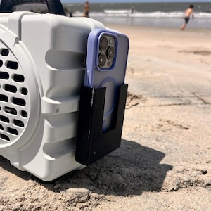 May include: A white portable speaker with a black handle and a black phone holder attached. A light purple phone case is in the holder. The speaker is on a sandy beach with the ocean in the background.