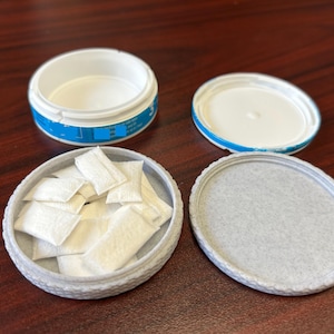 May include: An open, round container with a white interior and a blue and white label. The container is filled with small, white pouches. The lid is next to the container. The container and lid are on a dark brown surface.