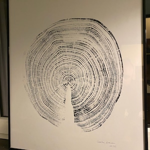 Op de afbeelding: Zwart-wit ingelijste print van een dwarsdoorsnede van een boom, met jaarringen. Het kunstwerk, gesigneerd door Sacha Graven, is gedateerd 01/22. De print staat tegen een witte achtergrond, omlijst door een zwarte rand.