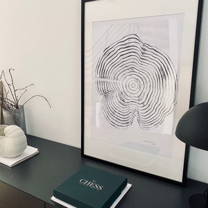 Op de afbeelding: Een zwart ingelijste print van een zwart-witte boomringontwerp op een witte achtergrond. De print ligt op een donkergrijs oppervlak met een groene doos waarop "No. 1 Chess" staat.