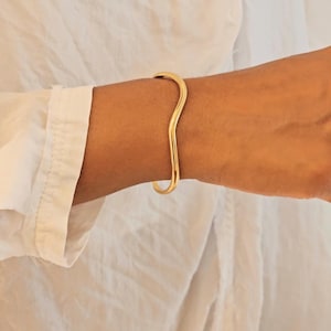 May include: A gold-colored, wavy cuff bracelet. The bracelet has a smooth, polished finish and a unique, organic shape. The jewelry is worn on a wrist, and the background is a white shirt.