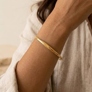 Pode incluir: Uma pulseira tipo bracelete de ouro polido e liso com um design de banda arredondada e contínua. O metal tem um acabamento brilhante e reflexivo que capta a luz, criando uma aparência elegante e uniforme ao redor do pulso.
