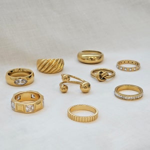May include: A collection of gold-toned rings with various designs, including a ring with a clear stone, a ring with a knot design, and a ring with a textured band.
