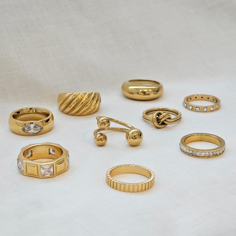 May include: A collection of gold-toned rings with various designs, including a ring with a clear stone, a ring with a knot design, and a ring with a textured band.