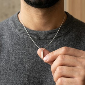Puede incluir: Un collar plateado con un colgante rectangular. El colgante cuelga de una cadena delicada. La persona de la imagen lleva un jersey gris. El collar tiene un diseño minimalista.