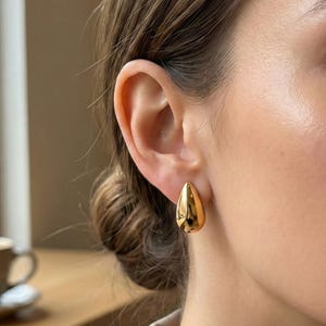 May include: A close-up of a person's ear with a pair of gold teardrop-shaped stud earrings. The earrings have a smooth, polished surface and reflect light, creating a shiny appearance. The earrings are a simple yet elegant accessory.