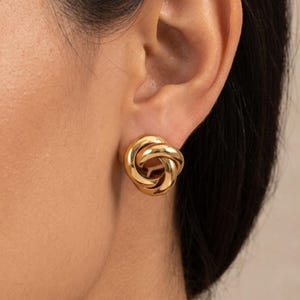 May include: Gold-colored knot stud earrings. The earrings are round and feature a three-dimensional knot design. The earrings are shown on an ear.