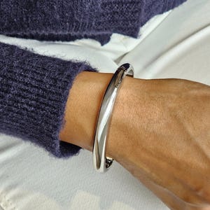 May include: A polished silver bangle. The bracelet is a solid, curved band with a smooth, reflective surface. The bangle is worn on a wrist, with a dark blue jumper in the background.