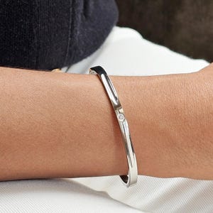 Puede incluir: Un brazalete de plata con un pequeño diamante redondo en el centro. El brazalete es liso y pulido, reflejando la luz. El brazalete se lleva en un brazo.