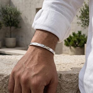 May include: A silver cuff bracelet with a textured pattern. The bracelet is worn on a wrist, and the person is wearing a white long-sleeved shirt. The bracelet appears to be made of metal and has a polished finish.