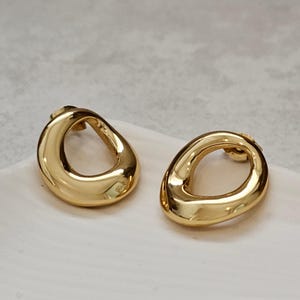 Pode incluir: Um par de brincos de pressão ovais abertos dourados. Os brincos têm uma superfície lisa e polida e um fecho de pino. Os brincos são exibidos em uma superfície branca.