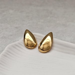 Könnte beinhalten: Ein Paar goldfarbene Ohrringe in Tropfenform. Die Ohrringe haben eine glatte, polierte Oberfläche, die das Licht reflektiert. Sie werden auf einer weißen, strukturierten Oberfläche präsentiert.
