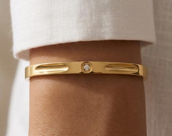 Puede incluir: Un brazalete rígido de tono dorado pulido con una banda lisa y redondeada que presenta un engaste circular central. Este engaste contiene una pequeña piedra preciosa transparente de talla redonda. La banda tiene dos hendiduras cóncavas.