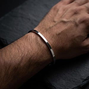 Può includere: Un bracciale a polsino in argento lucido. Il bracciale ha un design minimalista con una superficie liscia e riflettente. Il gioiello è indossato su un polso su uno sfondo scuro.