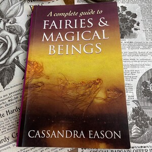 Könnte beinhalten: Ein Buch mit dem Titel "A complete guide to FAIRIES & MAGICAL BEINGS" von Cassandra Eason. Das Cover zeigt einen Farbverlauf von Gelb nach Lila mit Feenillustrationen. Das Buch liegt auf einem Hintergrund aus Vintage-Blumendrucken.