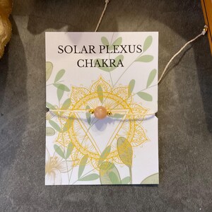 Op de afbeelding: Een armband met een perzik-kleurige stenen kraal en gouden accenten wordt getoond op een kaart. De kaart bevat de tekst "SOLAR PLEXUS CHAKRA" met een geel mandala-ontwerp en groene bladillustraties. De armband is geregen op een crèmekleurig koord.