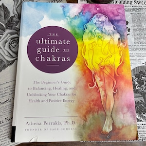 May include: A book titled "The Ultimate Guide to Chakras" with a watercolour illustration of a figure with flowing hair in shades of yellow, orange, and pink. The book cover has a purple circle with white text. The book is on a vintage newspaper.