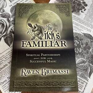 Könnte beinhalten: Ein Buch mit dem Titel "The Witch's Familiar" mit einer Vollmondgrafik. Das Cover zeigt eine Illustration einer Figur mit Besen und Schriftrolle. Das Buch ist von Vintage-Botanikausdrucken umgeben.