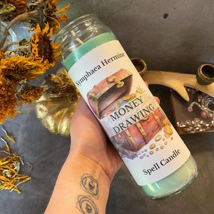 May include: A green spell candle with a label that reads "Money Drawing." The label features an illustration of a treasure chest overflowing with coins. The candle is in a clear glass jar.