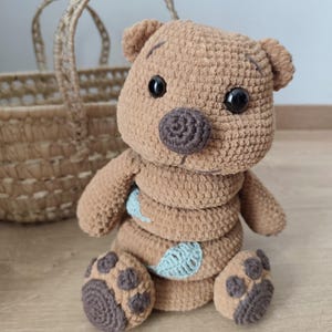 Peut inclure: Un ours en peluche au crochet marron fait à la main avec un design de corps empilé unique. L'ours a des yeux brun foncé, un nez rond et des accents de feuilles bleu clair. L'ours est assis sur une surface en bois.