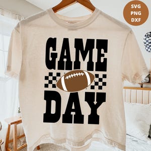 Peut inclure: Un t-shirt à manches courtes de couleur crème avec le texte noir "GAME DAY" dans une police grasse et carrée. Entre les mots se trouve un ballon de football américain marron et blanc, entouré de motifs à damier noir et blanc.