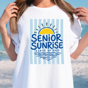 May include: White t-shirt with a blue and yellow graphic. The graphic features a yellow sun, blue text that reads "SENIOR SUNRISE" and "CLASS OF 2027", and blue water waves. The background has blue and white vertical stripes.