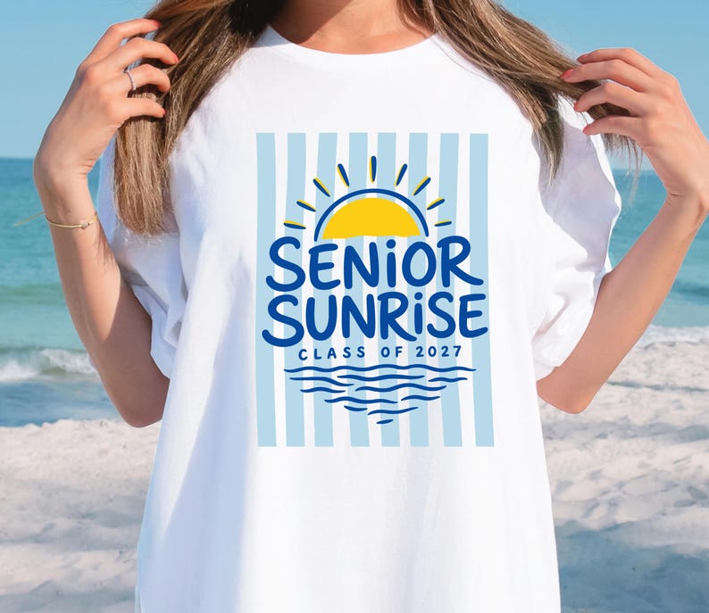 May include: White t-shirt with a blue and yellow graphic. The graphic features a yellow sun, blue text that reads "SENIOR SUNRISE" and "CLASS OF 2027", and blue water waves. The background has blue and white vertical stripes.