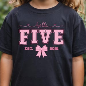Puede incluir: Camiseta negra con el texto rosa "hello FIVE", con un lazo rosa y el año "2021" debajo. También se incluye la palabra "EST.". La camiseta es probablemente para el quinto cumpleaños de un niño.