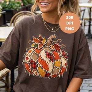Puede incluir: Una camiseta de manga corta de color marrón oscuro con un gran gráfico central de una calabaza construida con varios parches de tela texturizada en tonos naranja, rojo, amarillo y marrón, acentuada con patrones de hojas de otoño y cuadros.